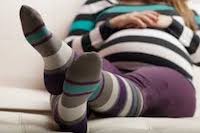 A pregnant woman wearing grey, blue, and purple striped compression socks while relaxing on her couch.