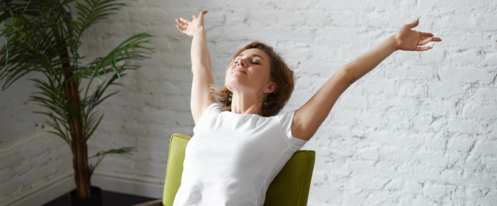 a woman with her arms outstretched takes a deep breath while seated with her back straight against a green chair