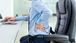 a seated man works at his computer in the office and seems to hold his lower back in a sign of pain