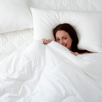 A smiling young woman lying in bed with a luxurious looking white duvet pulled up to her chin.